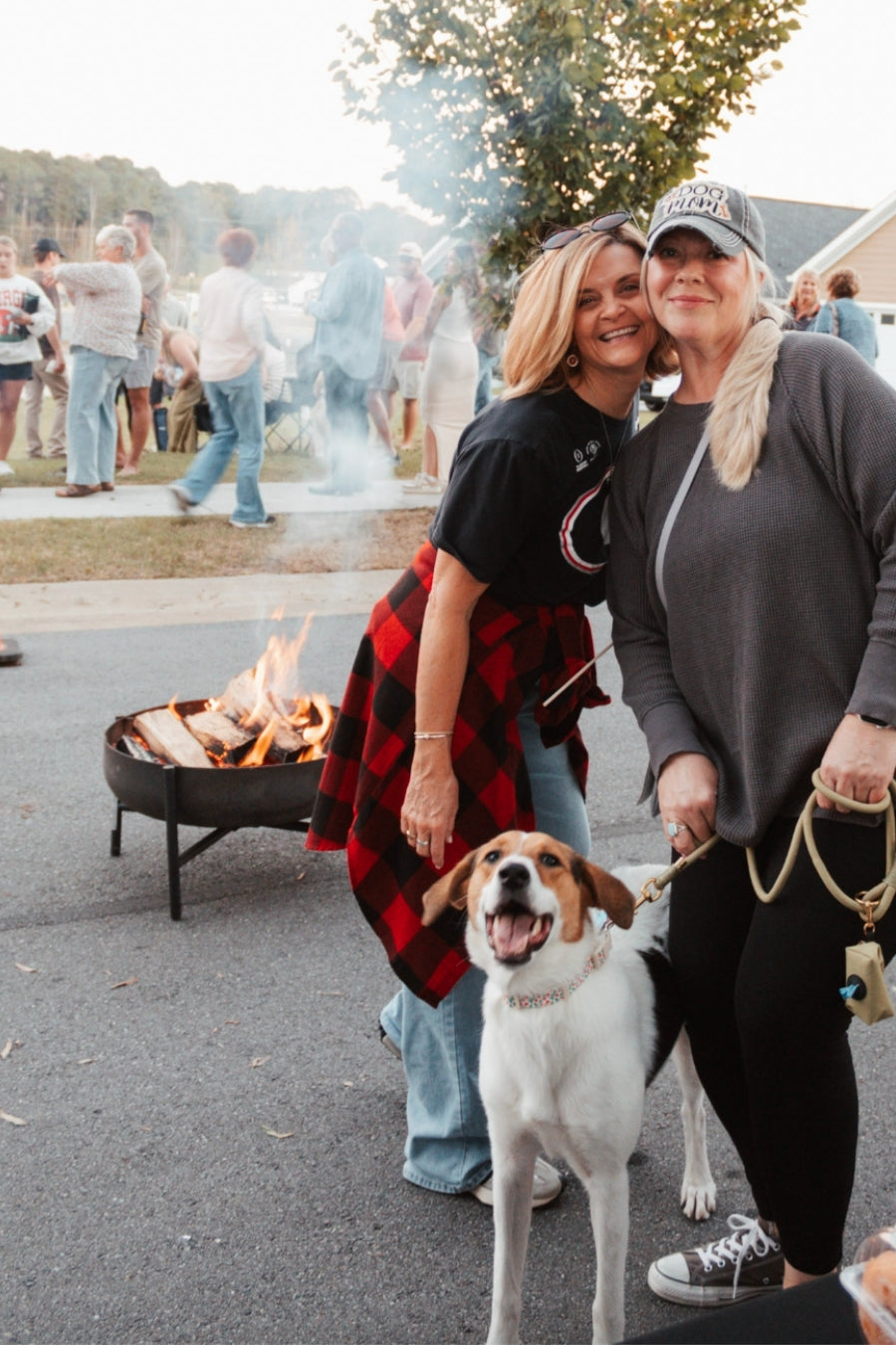 Two women and a dog standing in front of a fire pit with people and trees in the background. Dalton, GA