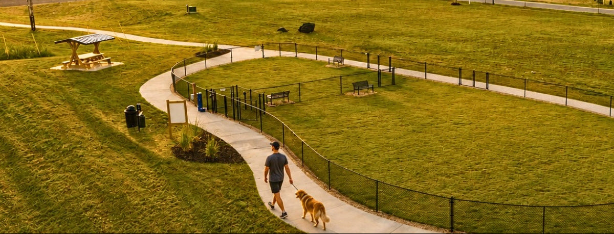 A wide outdoor park scene at sunset with a curved walking path, grassy fields, picnic tables, a fenced dog walk area, and a row of houses in the background. A person is walking a dog along the path near the bottom center.